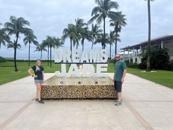 Two guests at the Dreams Jade resort sign with palm trees and Caribbean Sea in the background